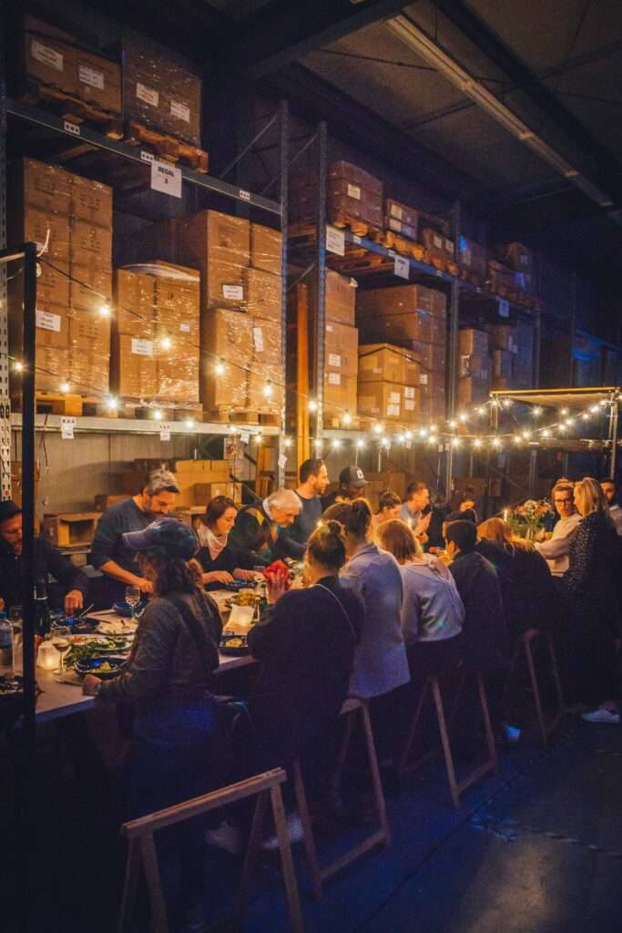 Gäste beim Dinner an der langen Tafel in einer Lagerhalle unter Lichterketten