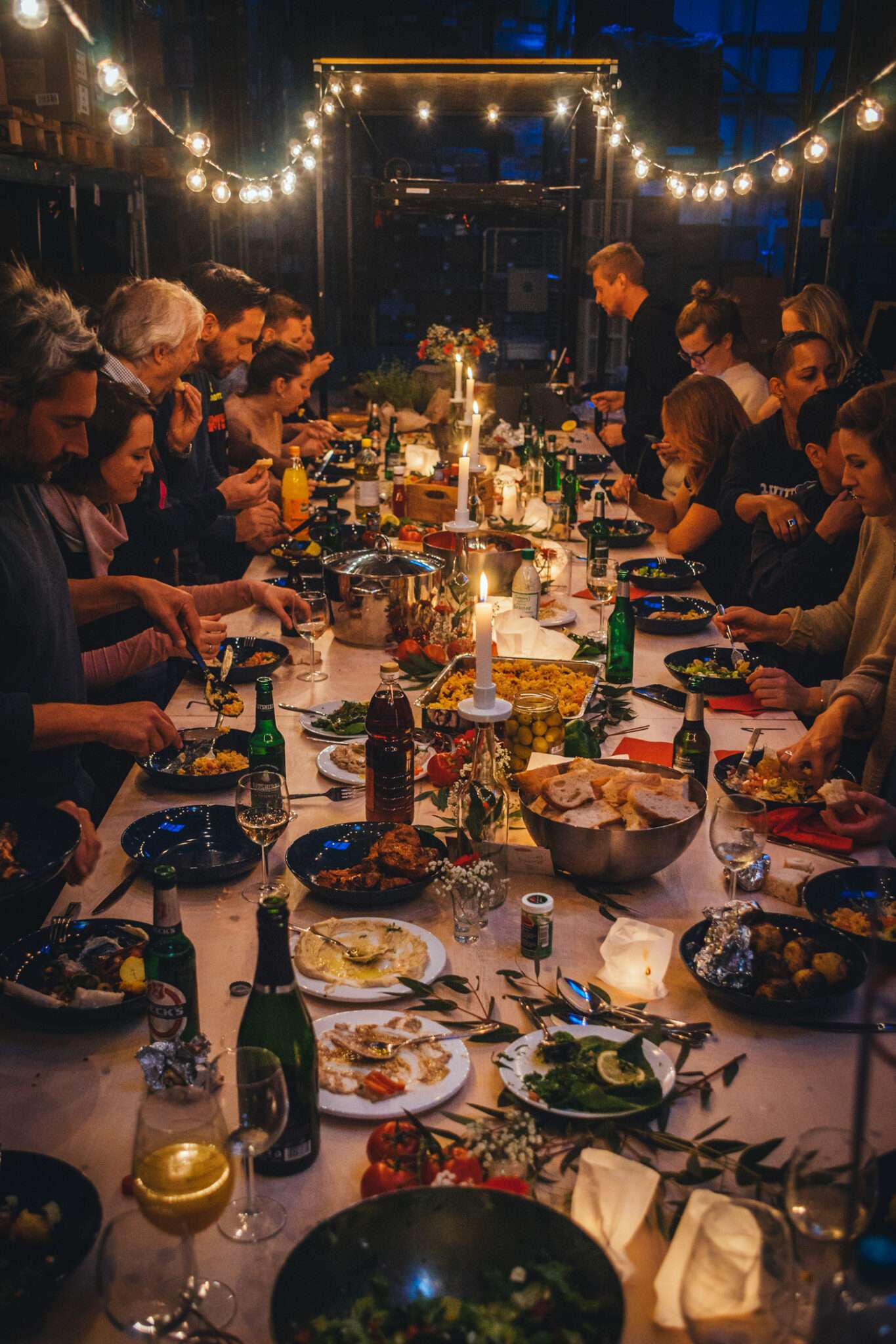 Lange Tafel mit vielen Gästen beim Dinner in einer Lagerhalle, Kerzen und Lichterketten