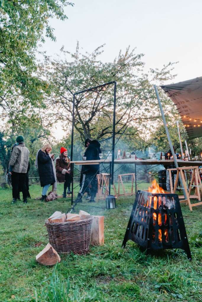 Feuertonne mit Brennholzkorb vor der langen Tafel unter Lichterketten im Grünen