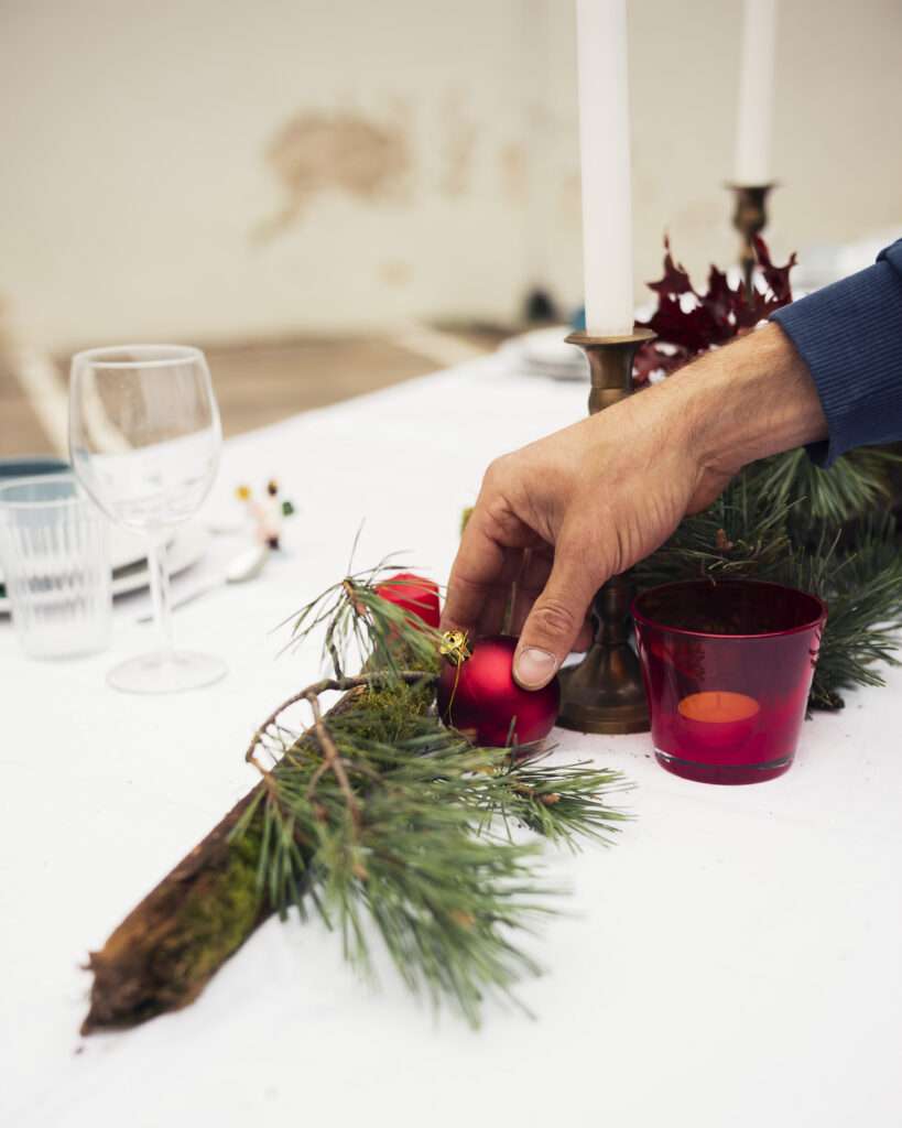 Hand legt eine rote Weihnachtskugel zwischen Tannenzweige auf der festlichen Tafel