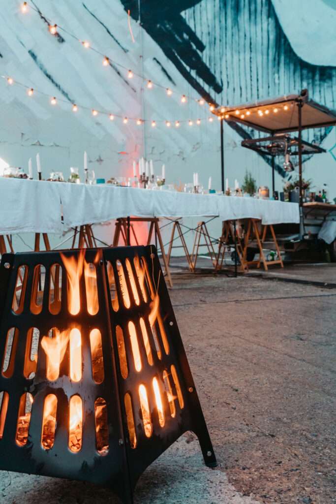 Brennende Feuertonne vor einer langen, festlich gedeckten Tafel mit Lichterketten